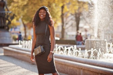 Şık Afro-Amerikan Kız gri tunik, crossbody çanta ve kap çeşmeler karşı güneşli sonbahar günü, poz verdi. Afrika model kadın.