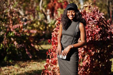 Şık Afro-Amerikan Kız gri tunik, crossbody çanta ve kapağı kırmızı yaprakları karşı güneşli sonbahar günü, poz verdi. Afrika model kadın.