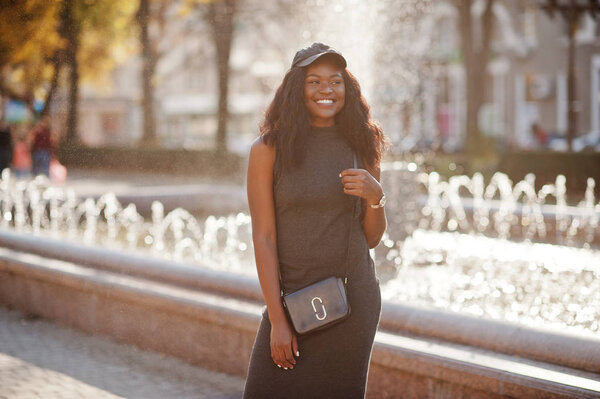 Стильная африканская американская девушка в серой тунике, мешок для перекрещивания тела и кепка позировали в солнечный осенний день против фонтанов. Африканская модель женщины
.