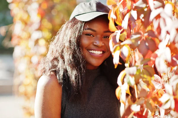 Stylish african american girl in cap posed at sunny autumn day against red leaves. Africa model woman.