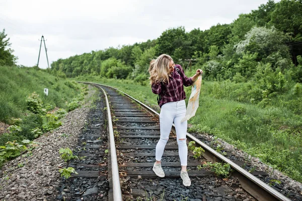 Portrait of a pretty blond girl in tartan shirt walking on the railway with map in her hands.