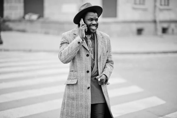 Stylish African American man model in gray coat, jacket tie and red hat walking on crosswalk and speaking on mobile phone. Black and white photo.