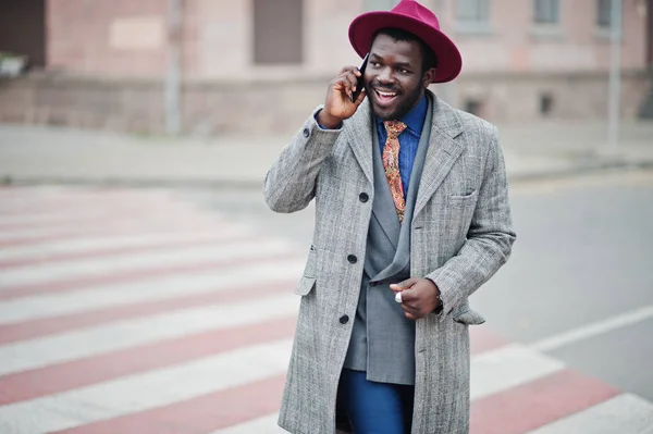 Stylish African American man model in gray coat, jacket tie and red hat walking on crosswalk and speaking on mobile phone.