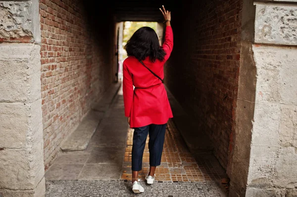 Back of stylish african american woman in red coat posed at tonel outdoor and shows two fingers sign.