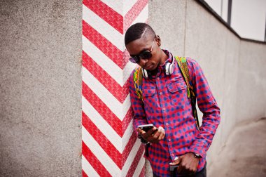 Afrika kökenli Amerikalı adam kareli gömlek, güneş gözlüğü ve kulaklık. Siyah adam gezgin cep telefonu tutan duvara.