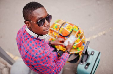 Kareli gömlek, güneş gözlüğü ve earphones ile bavul ve sırt çantası bir Afrika kökenli Amerikalı adam. Cep telefonu el altında tutarak siyah adam traveler.