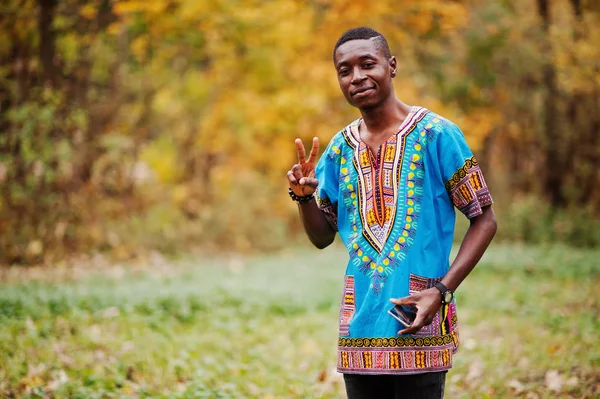 African man in africa traditional shirt on autumn park shows two fingers sign.