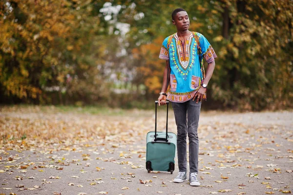 African man in africa traditional shirt on autumn park with backpack and suitcase. Emigrant traveler.