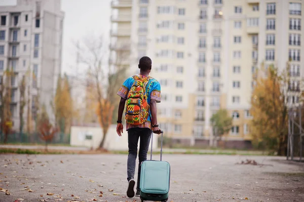 African man in africa traditional shirt on autumn park with backpack and suitcase. Emigrant traveler.