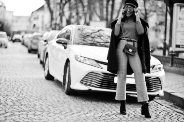 Afro-Amerikan moda kız ceket, haber teIIaIı şapka ve güneş gözlüğü Street beyaz iş araba karşı poz verdi.