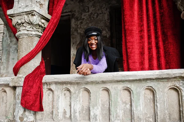 African american woman at violet dress and cap posed outdoor against old column with red curtains.