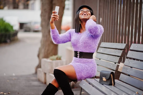 African american woman at violet dress and cap posed outdoor, sitting on bench and making selfie on phone.