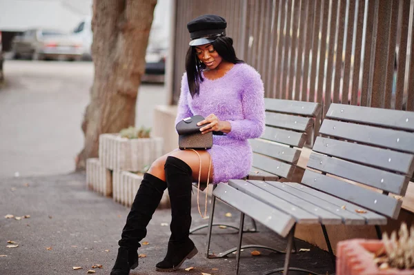 African american woman at violet dress and cap posed outdoor, sitting on bench and finding something at her handbag.