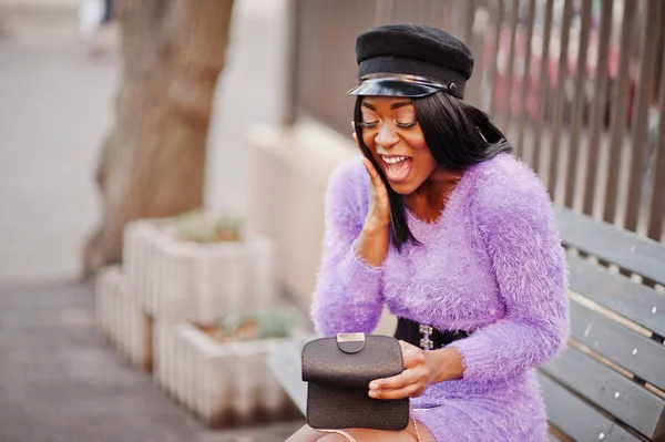 African american woman at violet dress and cap posed outdoor, sitting on bench and finding something at her handbag.
