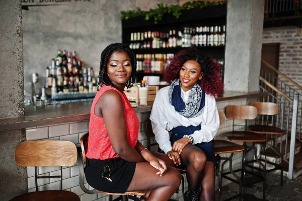 Attractive african american two girls friends sitting at chairs against bar on pub.