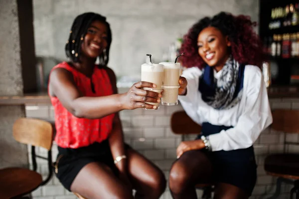 Attractive african american two girls friends sitting at chairs against bar on pub and drinking latte.