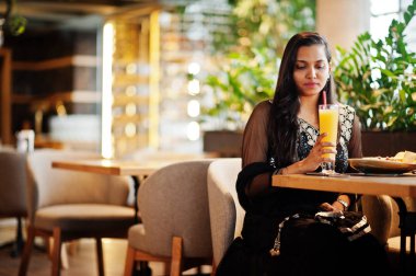 Restoranda, suyu ve salata ile masada oturan siyah sari giysili güzel Hintli kız poz.