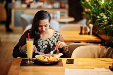 Restoranda, suyu ve salata ile masada oturan siyah sari giysili güzel Hintli kız poz.