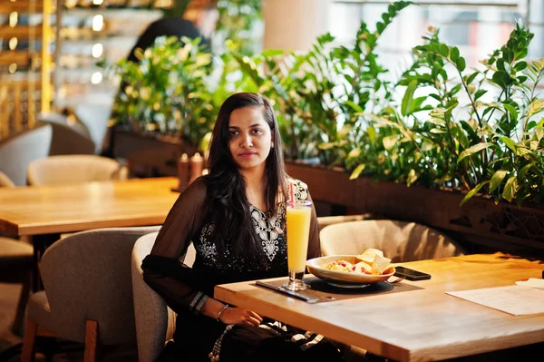 Restoranda, suyu ve salata ile masada oturan siyah sari giysili güzel Hintli kız poz.