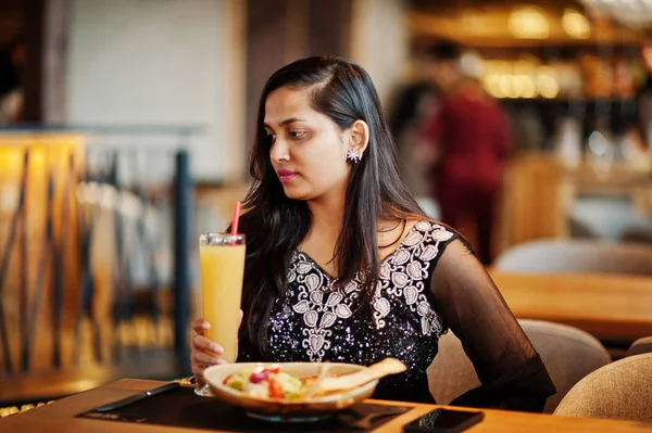 Restoranda, suyu ve salata ile masada oturan siyah sari giysili güzel Hintli kız poz.