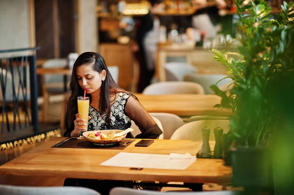Restoranda, suyu ve salata ile masada oturan siyah sari giysili güzel Hintli kız poz.