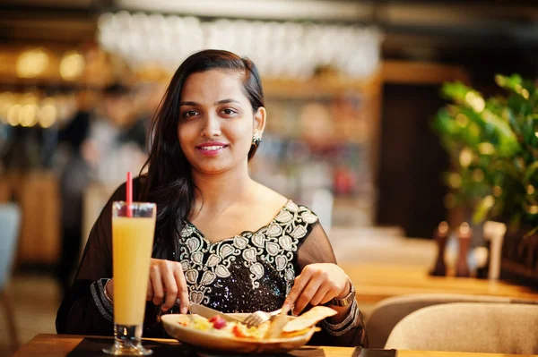 Restoranda, suyu ve salata ile masada oturan siyah sari giysili güzel Hintli kız poz.