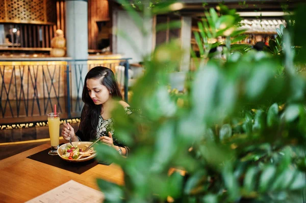 Restoranda, suyu ve salata ile masada oturan siyah sari giysili güzel Hintli kız poz.