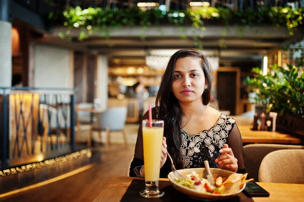 Restoranda, suyu ve salata ile masada oturan siyah sari giysili güzel Hintli kız poz.