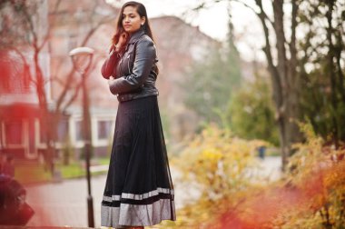 Pretty indian girl in black saree dress and leather jacket posed outdoor at autumn street. clipart