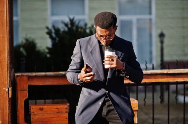 İnanılmaz Afro-Amerikan erkek giyimi mavi ceket broş ile seyir, siyah boğazlı kazak ve gözlük Sokağı'nda poz verdi. Fincan kahve ve cep telefonu şık siyah adam.