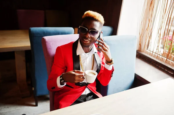 Fashion african american man model at red suit, with highlights hair and sunglasses sitting at cafe with coffee and speaking on mobile phone.