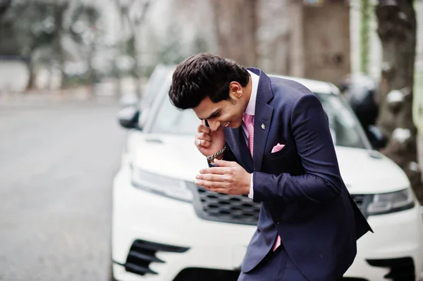 Elegant indian macho man model on suit and pink tie posed against white business car and speaking on phone.