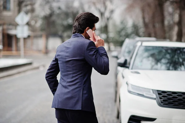 Elegant indian macho man model on suit and pink tie posed against white business car and speaking on phone.