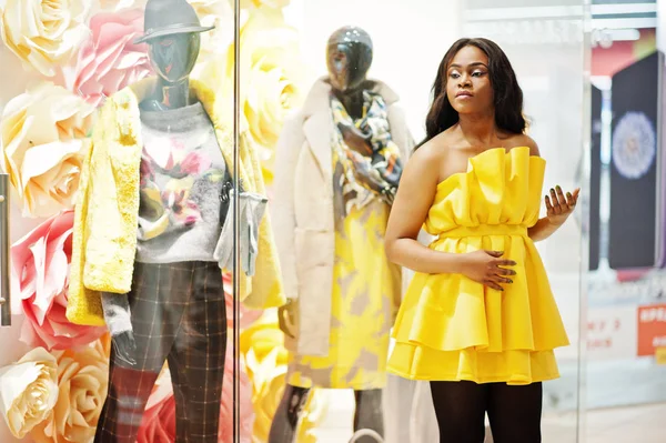 Stylish african american woman at yellow dreess posed against showcases with mannequins at mall.