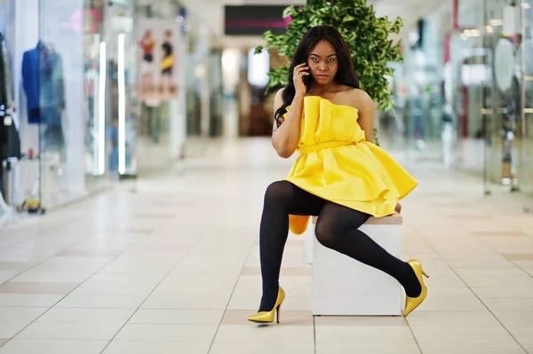 Stylish african american woman at yellow dreess posed at mall and sitting on bench inside, speaking at phone.