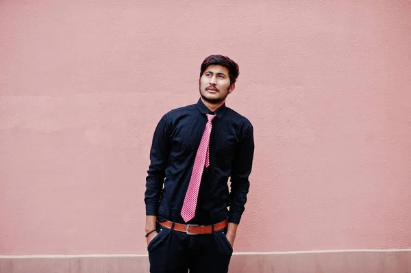 Young indian man on black shirt and tie posed against pink wall.
