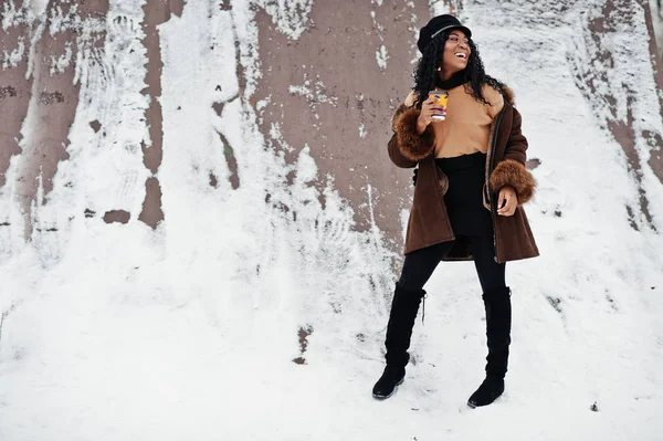 ムートン コートとキャップのアフリカ系アメリカ人の女性が一杯のコーヒーで雪に覆われた背景に冬の日で提起されました — ストック写真
