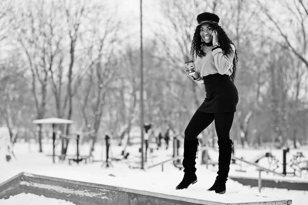 一杯のコーヒーを保持していると 電話で話す 雪に覆われた背景には 冬の日に黒スカート 茶色のセーターとキャップのアフリカ系アメリカ人の女性がもたらされる — ストック写真