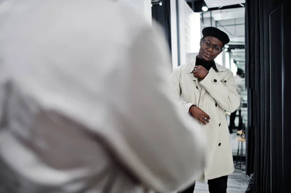 Stylish casual african american man at black beret and white trench raincoat at fitting room clothes store , looking at mirror.