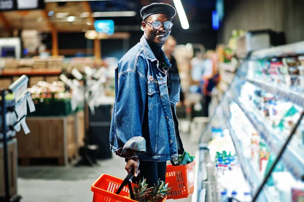 Stylish casual african american man at jeans jacket and black beret holding two baskets, standing near fridge and shopping at supermarket.