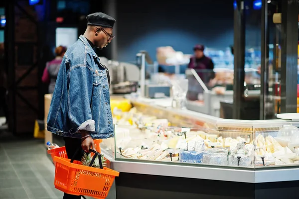 Stylish casual african american man at jeans jacket and black beret holding basket, standing near cheese fridge and shopping at supermarket.