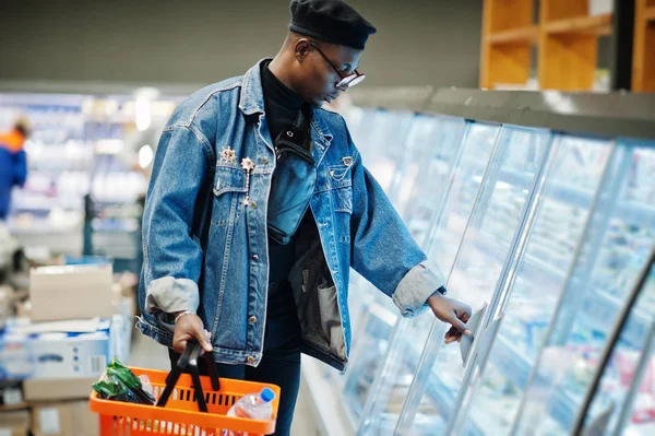 Stylish casual african american man at jeans jacket and black beret holding basket, standing near gastronomic fridge and shopping at supermarket.