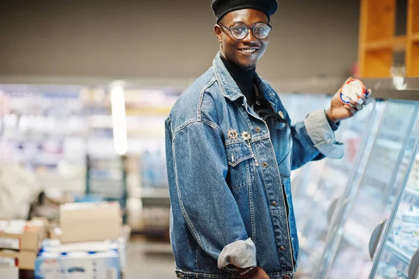 Stylish casual african american man at jeans jacket and black beret holding basket, standing near gastronomic fridge and shopping at supermarket.