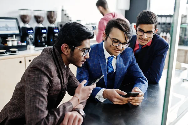 Group of three indian businessman in suits sitting on cafe and looking on mobile phone.