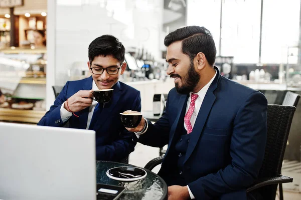 Two indian business man in suits sitting at office on cafe, looking at laptop and drinking coffee.