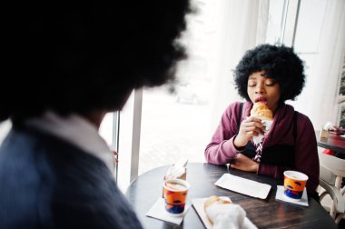 Afrikalı Amerikan Bayan giyim kazak üzerinde tablo kafede oturur iki kıvırcık saçlı kruvasan ve içecek çay yemek.