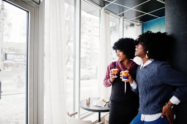Two curly hair african american woman wear on sweaters with cups of tea posed at cafe indoor.