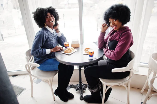 Two curly hair african american woman wear on sweaters sits at table cafe , eat croissant and drink tea.