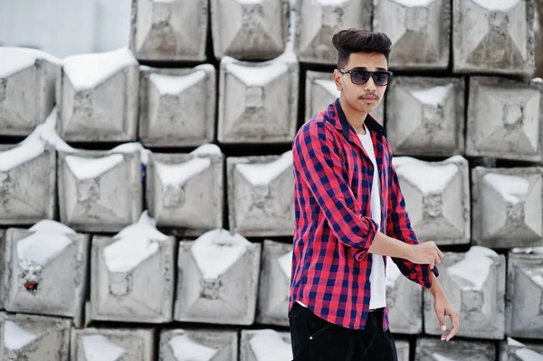 Casual young indian man in checkered shirt and sunglasses posed against stone blocks.