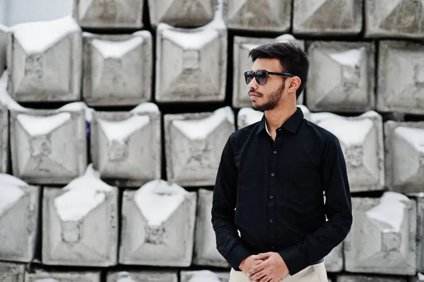 Casual young indian man in black shirt and sunglasses posed against stone blocks.